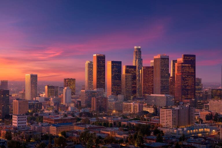 Downtown city skyline at sunset, with tall buildings reflecting warm sunlight under a colorful sky. Trees and smaller buildings are visible in the foreground, highlighting a vibrant landscape ideal for Independent Cyber Risk Diligence.